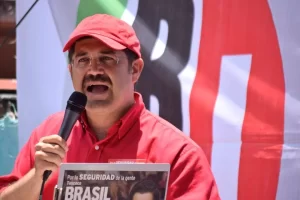 Hombre con camisa y gorra roja hablando con micrófono frente a pancarta