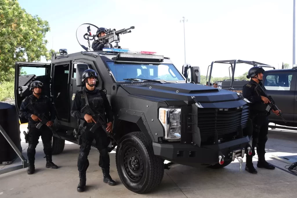 Cuatro policías uniformados con equipo táctico y armas, junto a un vehículo blindado negro