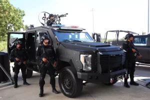 Cuatro policías uniformados con equipo táctico y armas, junto a un vehículo blindado negro