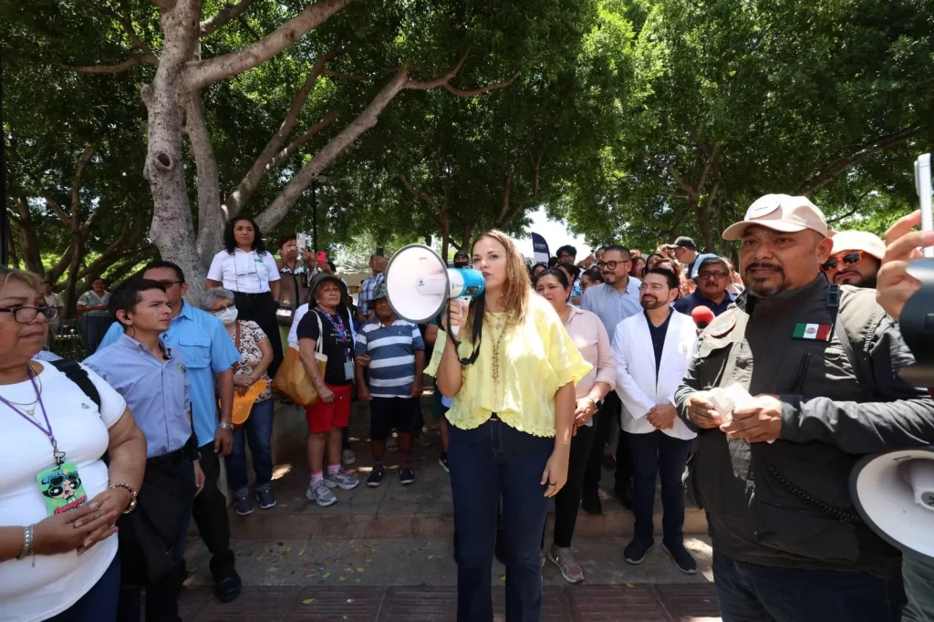 Mujer con blusa amarilla hablando por megáfono frente a grupo de personas bajo árboles