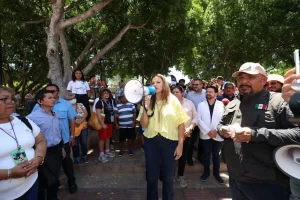 Mujer con blusa amarilla hablando por megáfono frente a grupo de personas bajo árboles