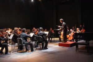Director de orquesta frente a músicos tocando violines y violonchelos en un escenario
