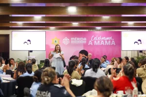 Mujer hablando con micrófono frente a grupo de personas en evento con banner rosa de 'Celebremos a mamá'