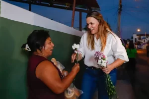 Mujer de cabello rubio entrega flores a señora que lleva pan en mano afuera de una casa al anochecer