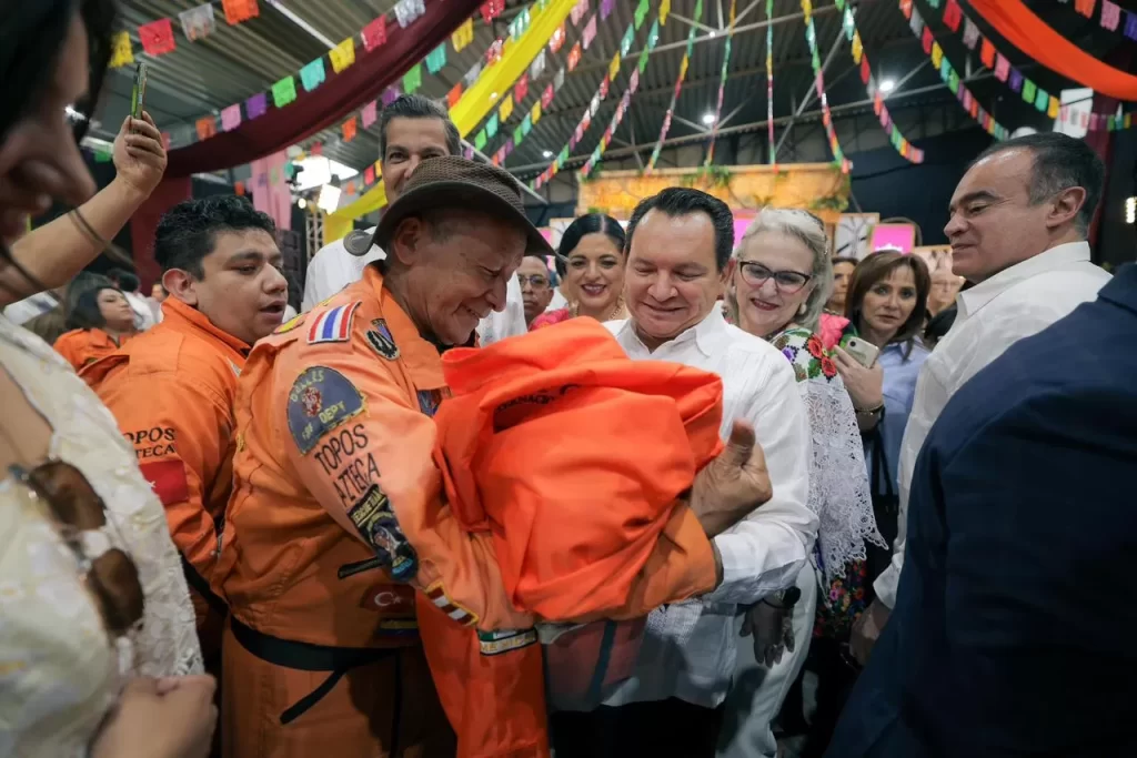 Hombre con chaqueta naranja de Topos Azteca recibiendo reconocimiento de grupo de personas en evento interior decorado con pa