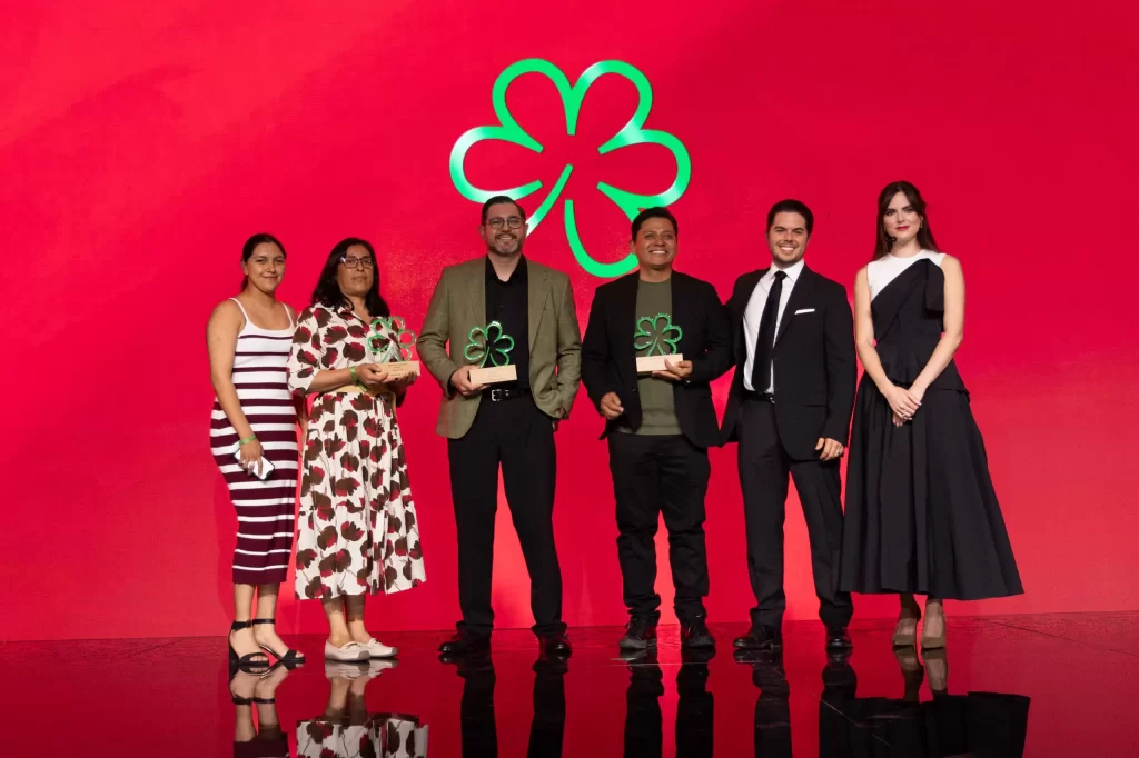 Seis personas en escenario con fondo rojo y trébol verde, tres con trofeos trébol