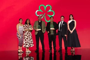 Seis personas en escenario con fondo rojo y trébol verde, tres con trofeos trébol