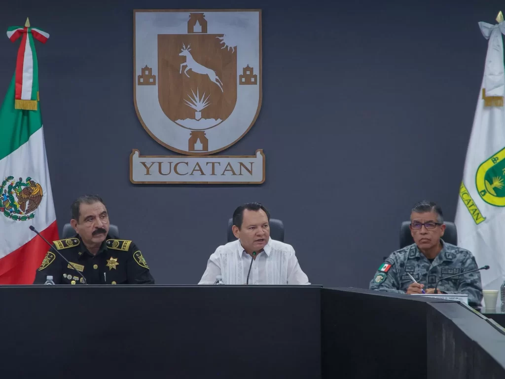 Tres hombres en una conferencia, uno con uniforme negro de policía, otro con guayabera blanca y otro con uniforme militar gri