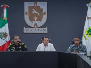 Tres hombres en una conferencia, uno con uniforme negro de policía, otro con guayabera blanca y otro con uniforme militar gri