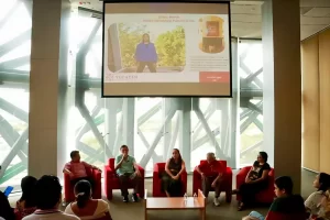 Cinco personas sentadas en sillones rojos participando en panel frente a audiencia, con proyección en pantalla grande