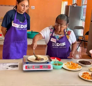 Dos mujeres con mandiles morados del DIF Yucatán preparando comida tradicional