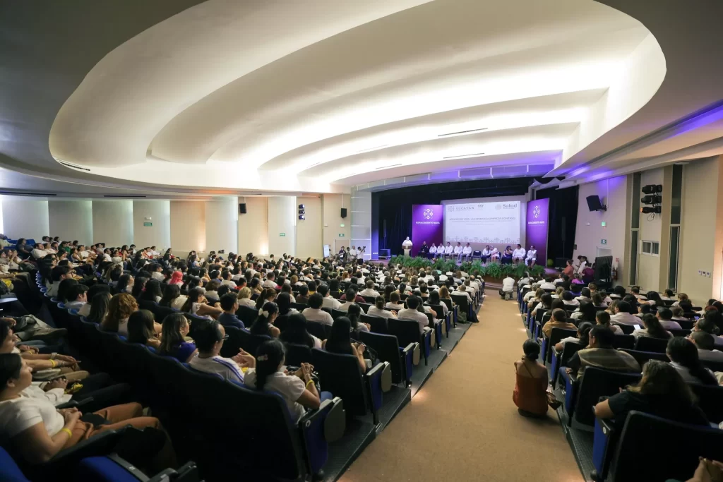 Personas sentadas en un auditorio escuchando una conferencia en escenario con pantalla y logos