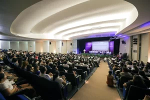 Personas sentadas en un auditorio escuchando una conferencia en escenario con pantalla y logos