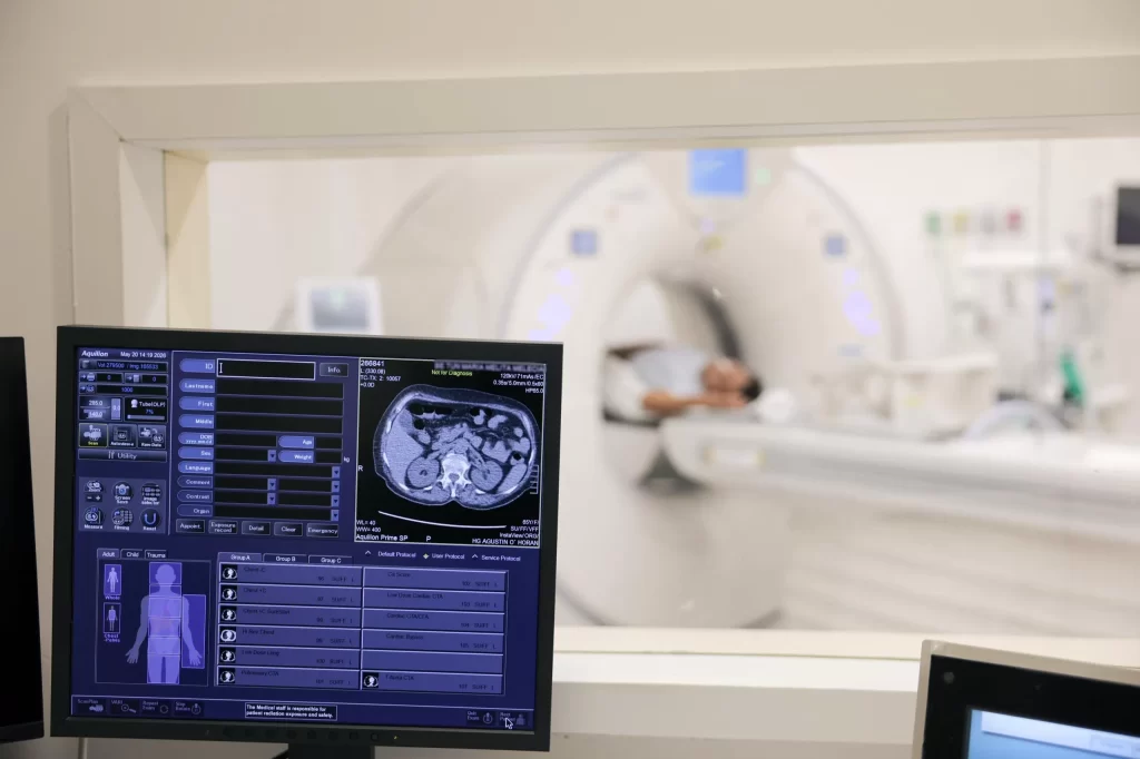 Pantalla mostrando imagen de tomografía axial de abdomen y controles médicos, con paciente en escáner en habitación contigua