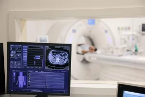 Pantalla mostrando imagen de tomografía axial de abdomen y controles médicos, con paciente en escáner en habitación contigua