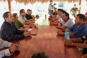 Personas sentadas a los lados de una mesa larga de madera en una reunión en un espacio con techo de palma.