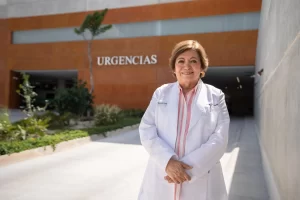 Mujer doctora con bata blanca sonriendo frente al acceso a urgencias en hospital