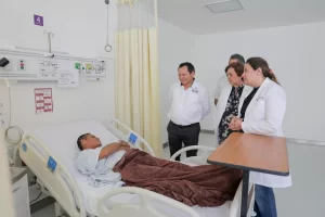 Paciente en cama de hospital con tres personas observando, una mujer con bata blanca