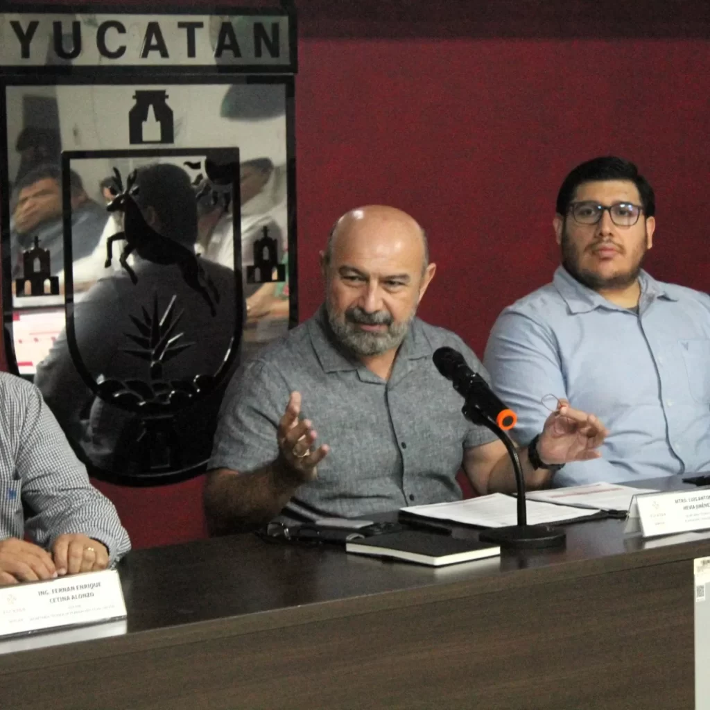 Tres hombres sentados en mesa con micrófono y escudo de Yucatán detrás