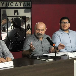 Tres hombres sentados en mesa con micrófono y escudo de Yucatán detrás