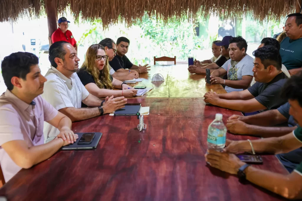 Varias personas sentadas en una mesa larga de madera bajo un techo de palma, en reunión o junta