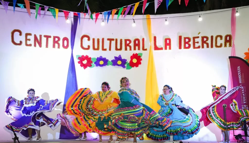 Mujeres bailando en vestidos típicos de colores vibrantes en el Centro Cultural La Ibérica decorado con papel picado y flores