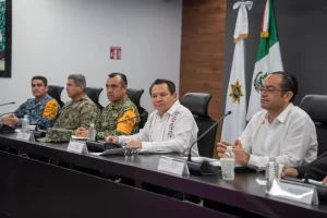 Cinco hombres sentados en mesa de conferencia en reunión formal; dos llevan uniformes militares con brazaletes naranjas y tre