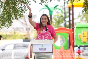 Mujer sonriente dando un discurso al aire libre frente a un podio con micrófono y logo de Mérida Enchula, parque infantil de