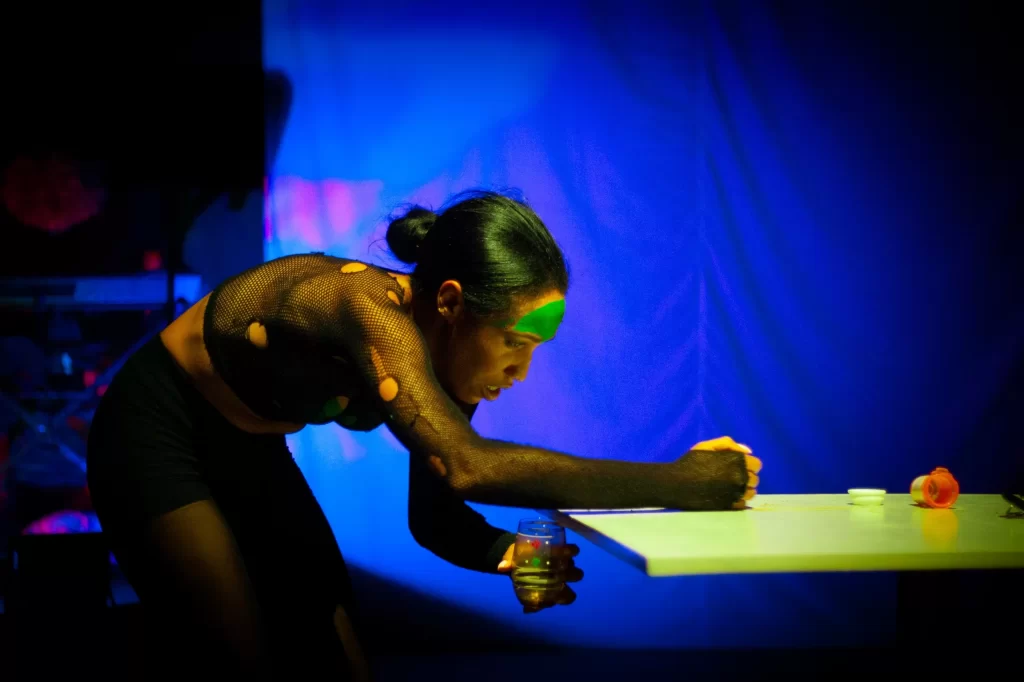 Persona con pintura verde en la cara y suéter de malla negra, inclinada sobre mesa blanca en ambiente con iluminación azul y