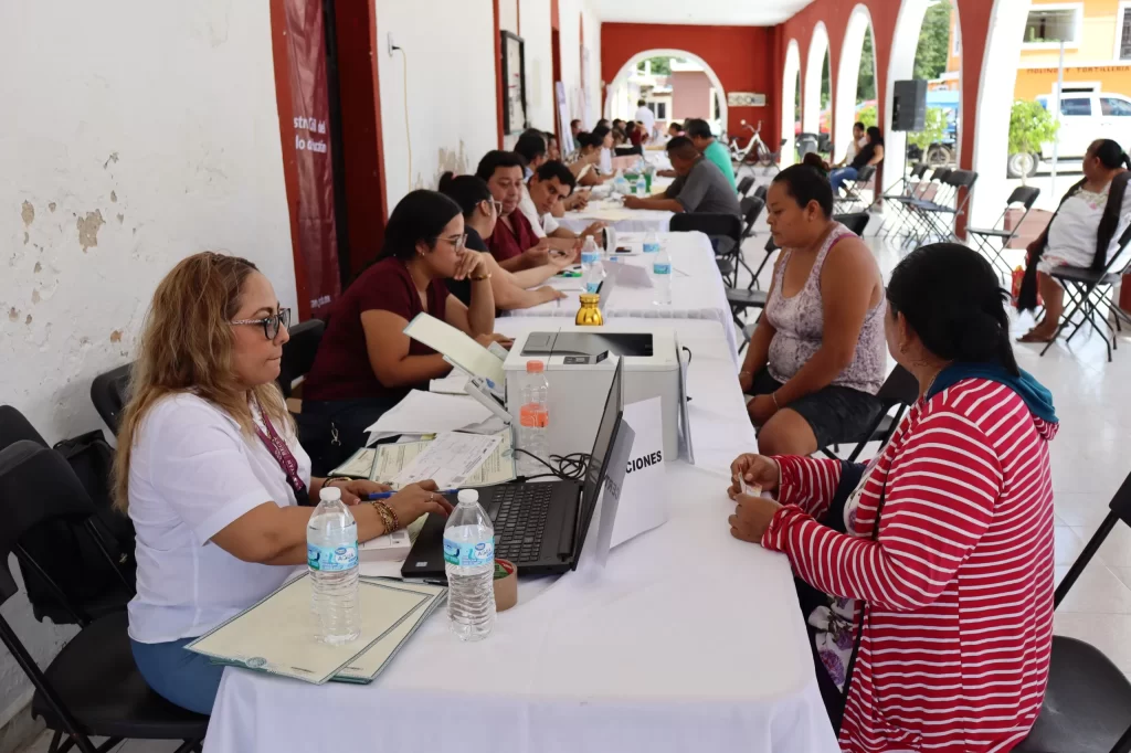 Personas sentadas en fila atendiendo trámites en mesa larga dentro de un recinto con arcos y paredes blancas