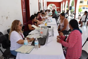 Personas sentadas en fila atendiendo trámites en mesa larga dentro de un recinto con arcos y paredes blancas