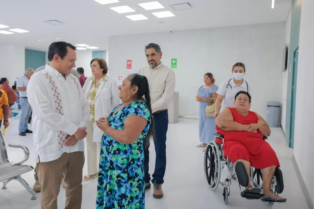 Hombre de camisa blanca hablando con mujer de vestido azul mientras otras personas esperan en área blanca de hospital