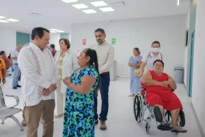 Hombre de camisa blanca hablando con mujer de vestido azul mientras otras personas esperan en área blanca de hospital