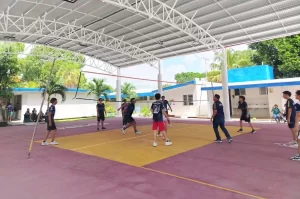 Jóvenes jugando voleibol en cancha techada con red y público al fondo