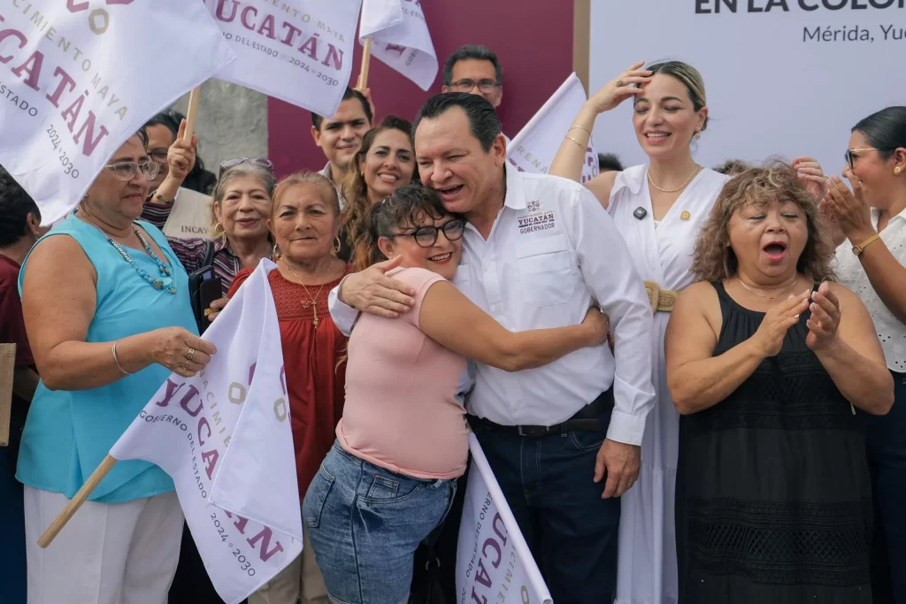 Gobernador de Yucatán abrazando a una mujer entre varias personas con banderas de Renacimiento Maya