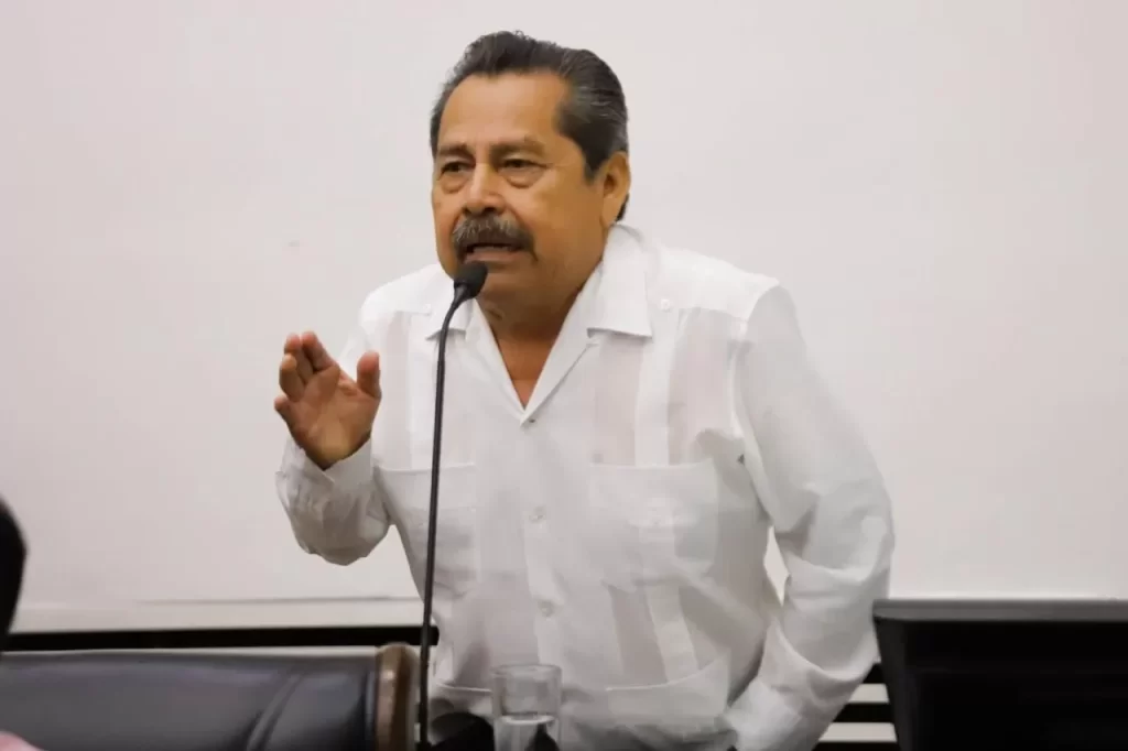 Hombre de mediana edad con camisa blanca de manga larga hablando frente a un micrófono con fondo blanco