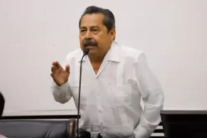 Hombre de mediana edad con camisa blanca de manga larga hablando frente a un micrófono con fondo blanco
