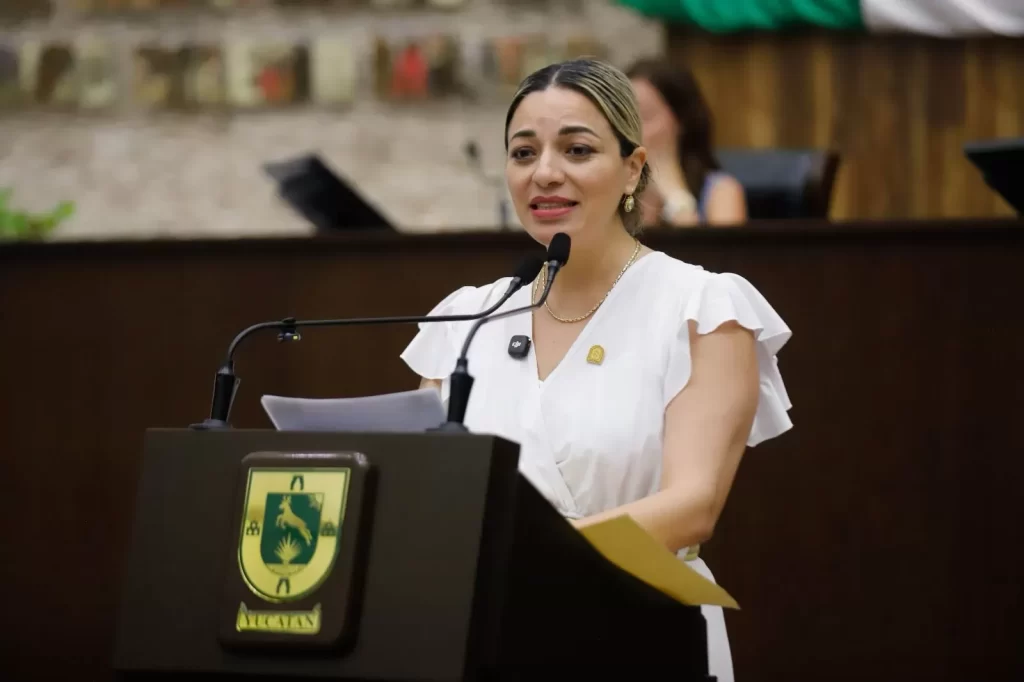 Mujer con blusa blanca hablando en un podium con dos micrófonos y el escudo de Yucatán