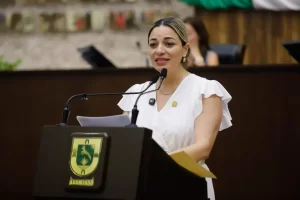 Mujer con blusa blanca hablando en un podium con dos micrófonos y el escudo de Yucatán
