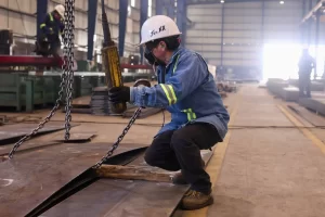 Hombre con casco blanco, chaleco reflectante y cubrebocas operando maquinaria en planta industrial
