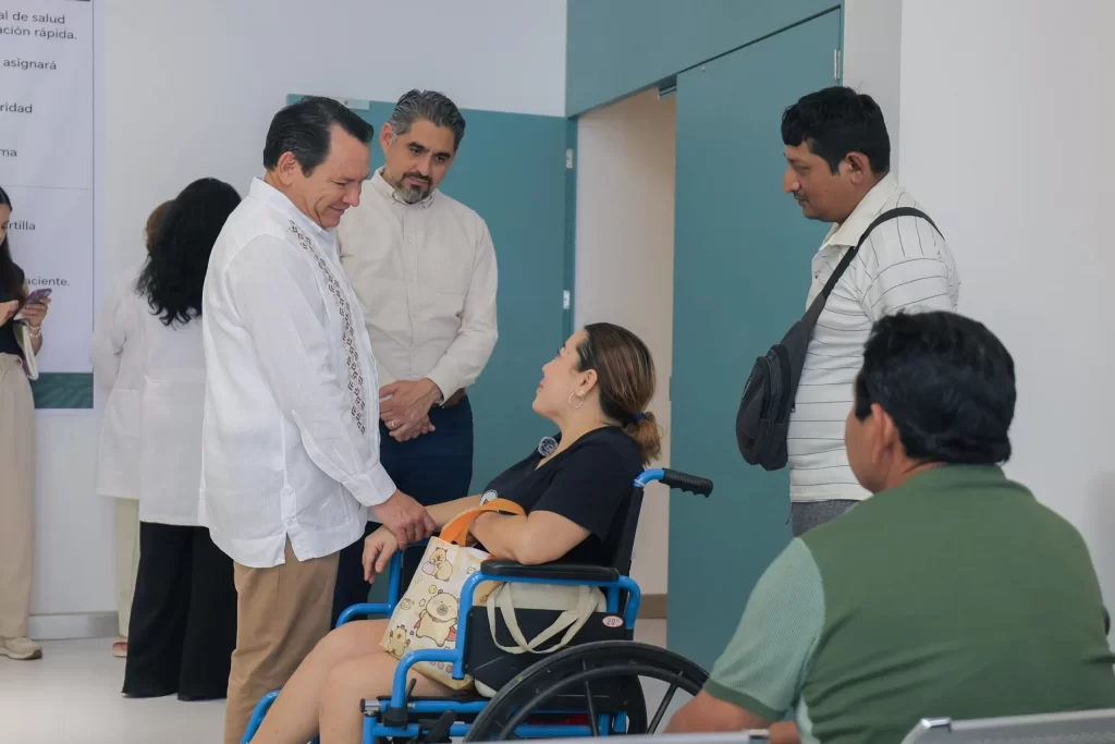 Mujer en silla de ruedas conversando con tres hombres en un pasillo de hospital, personas de pie y fondo con puerta azul