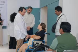 Mujer en silla de ruedas conversando con tres hombres en un pasillo de hospital, personas de pie y fondo con puerta azul