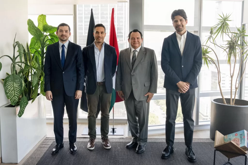 Cuatro hombres de traje y saco posan en oficina con plantas y dos banderas