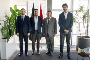 Cuatro hombres de traje y saco posan en oficina con plantas y dos banderas