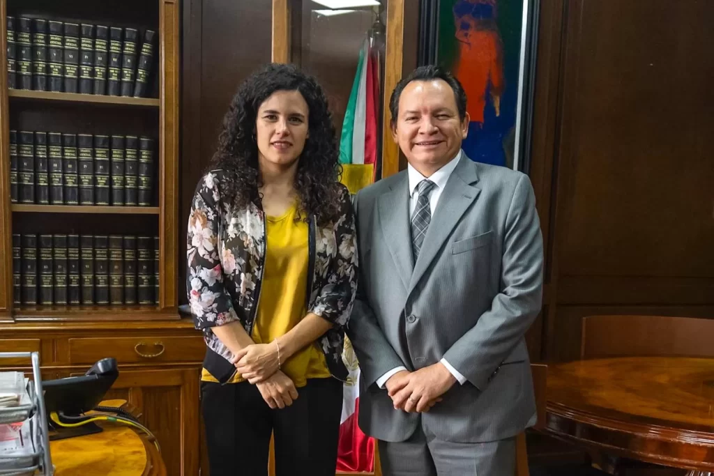 Dos personas posando en una oficina con estantería de libros y bandera mexicana