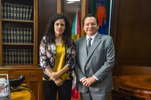Dos personas posando en una oficina con estantería de libros y bandera mexicana