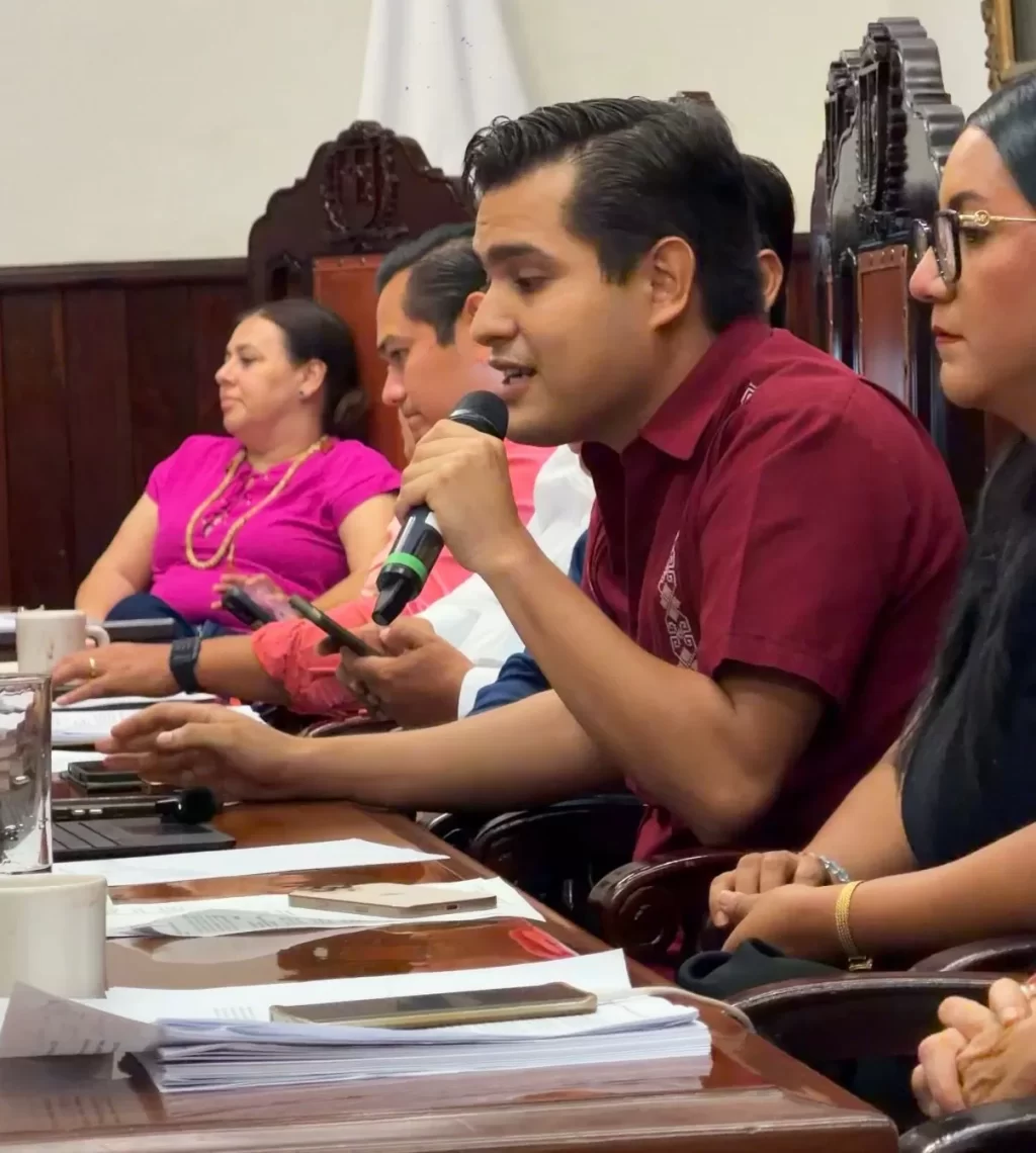 Hombre con camisa vino hablando con micrófono en mesa con documentos y personas a su alrededor