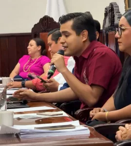 Hombre con camisa vino hablando con micrófono en mesa con documentos y personas a su alrededor