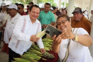 Hombre con guayabera blanca sostiene una mazorca de maíz blanco mientras una mujer con camiseta blanca se toma selfie con tel