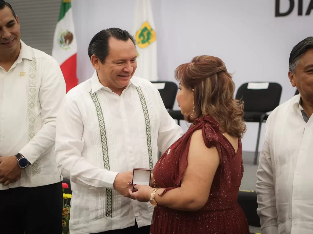 Hombre entregando un reconocimiento a una mujer en evento formal con banderas de México en el fondo